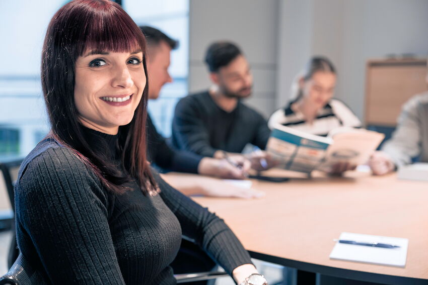 Eine MFORM-Mitarbeiterin lächelt in die Kamera, im Hintergrund sitzen Personen